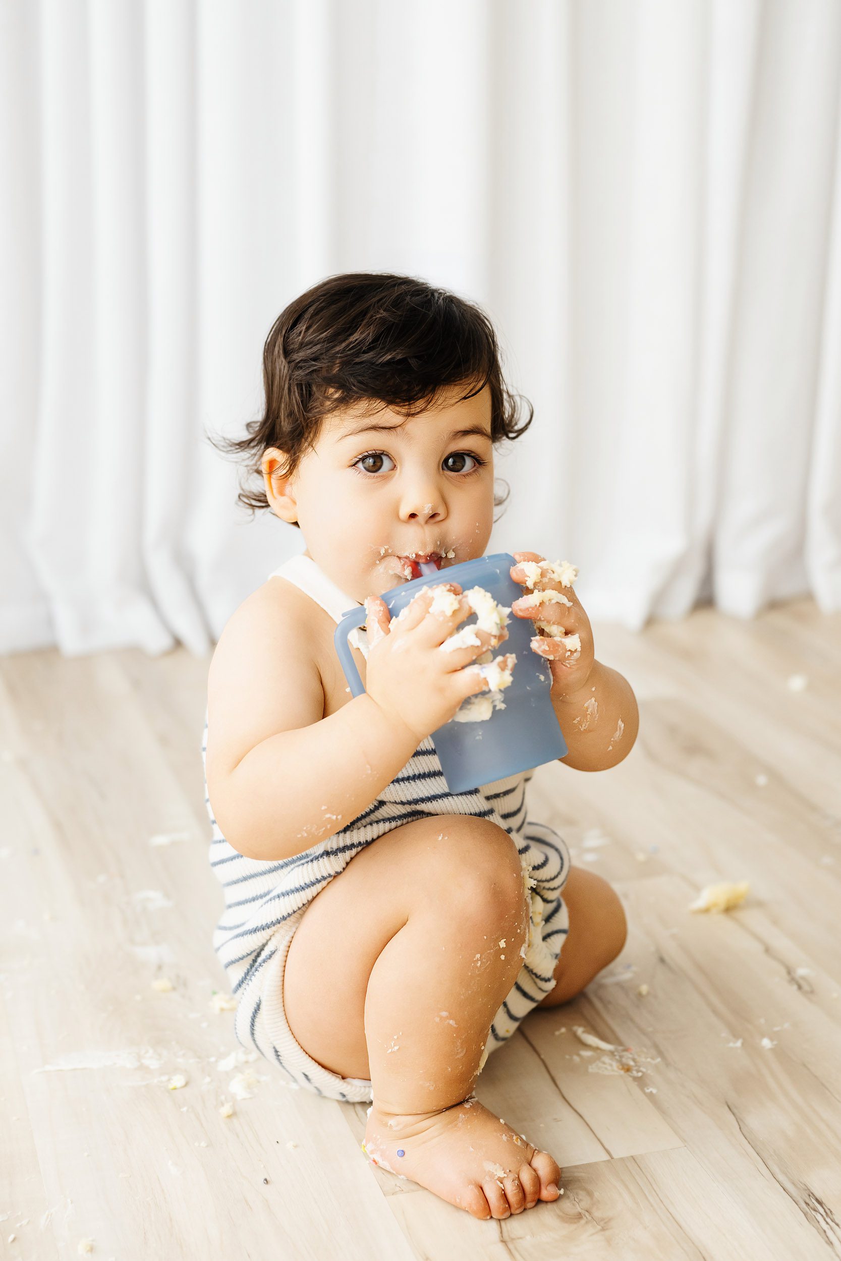 a young boy with cake and icing all of his arms, legs, hands, and face, taking a break from his cake smash photos for a drink from his water bottle as he gazes directly at the camera