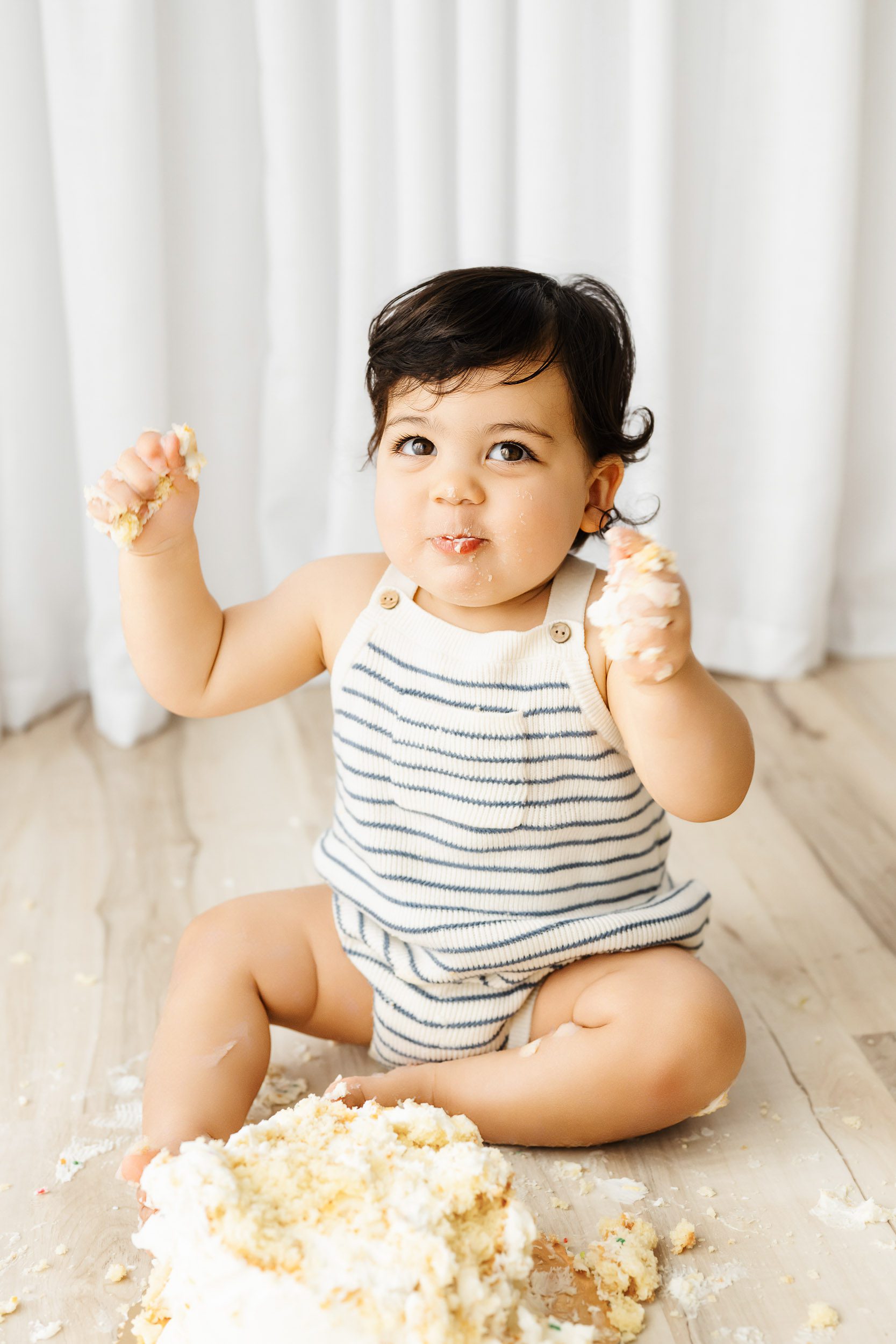 a little boy with a mouth full of cake grinning at the camera as he raises his arms in the air with his fists full of cake  during a cake smash photoshoot