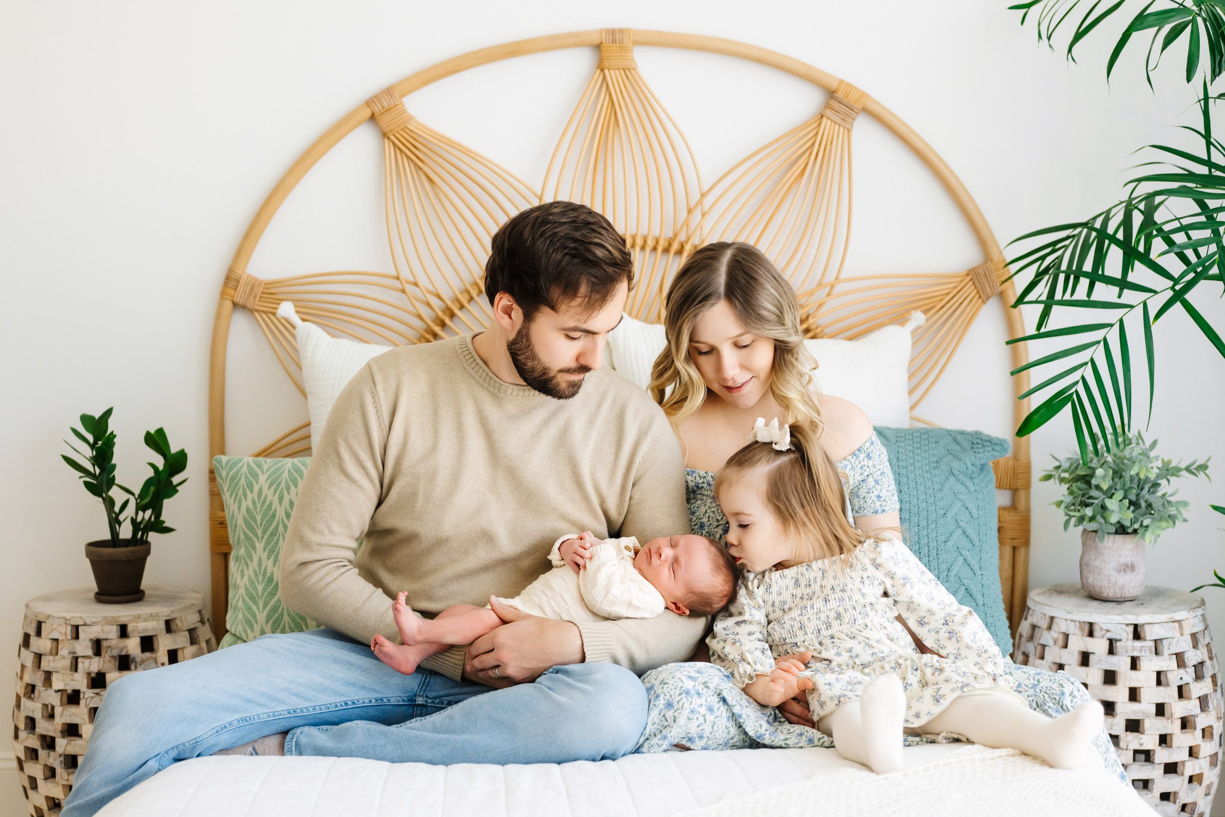 a family of four sitting on a bed with their baby boy cradled in dad's arms and big sister sitting on mom's lap as she leans over and gives her baby brother a kiss on his head during a newborn photography session near me