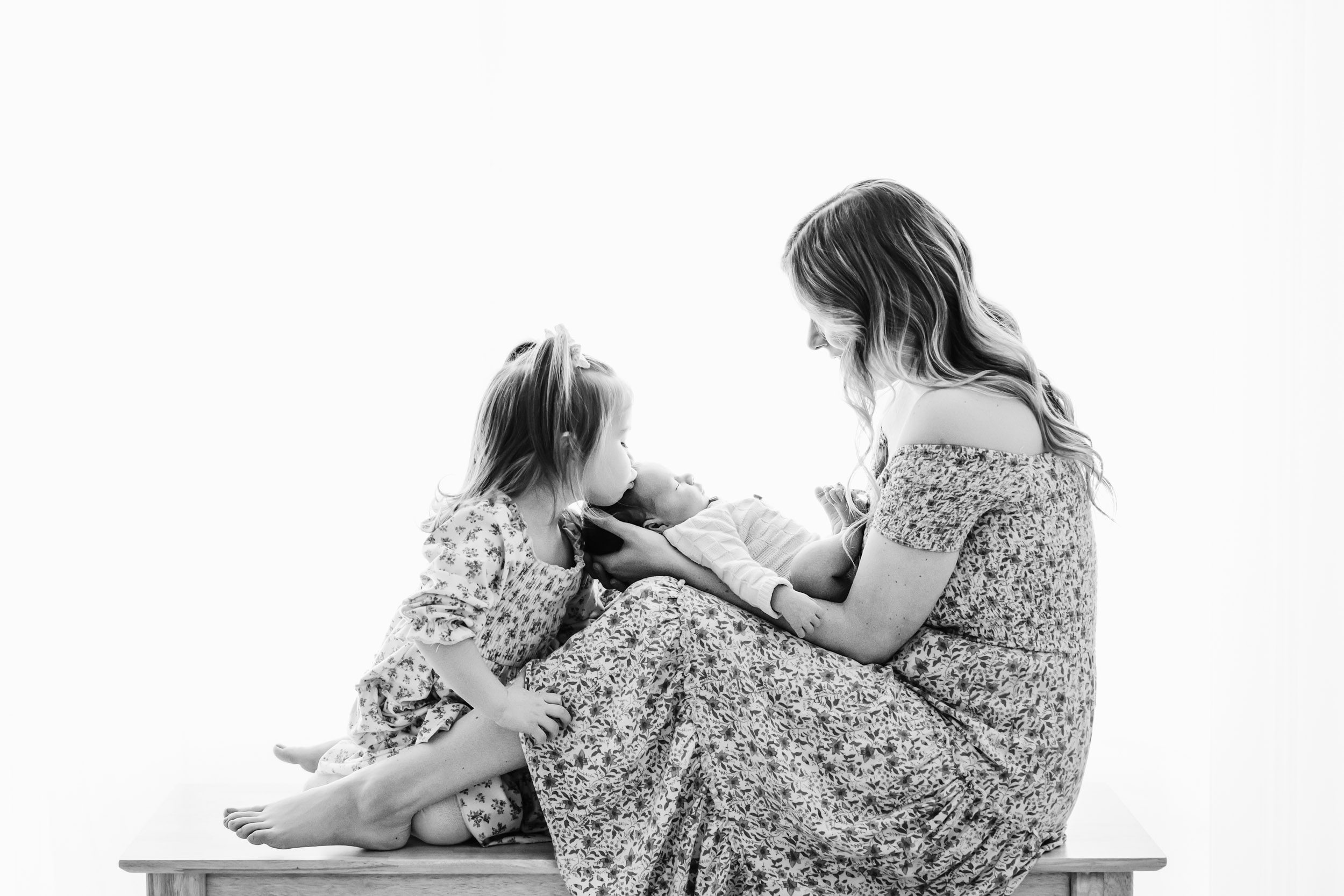 a black and white backlit photo of a mom sitting on a bench holding her baby boy on her legs as his big sister leans in and gives her baby brother a gentle kiss on his head during a newborn photography session near me