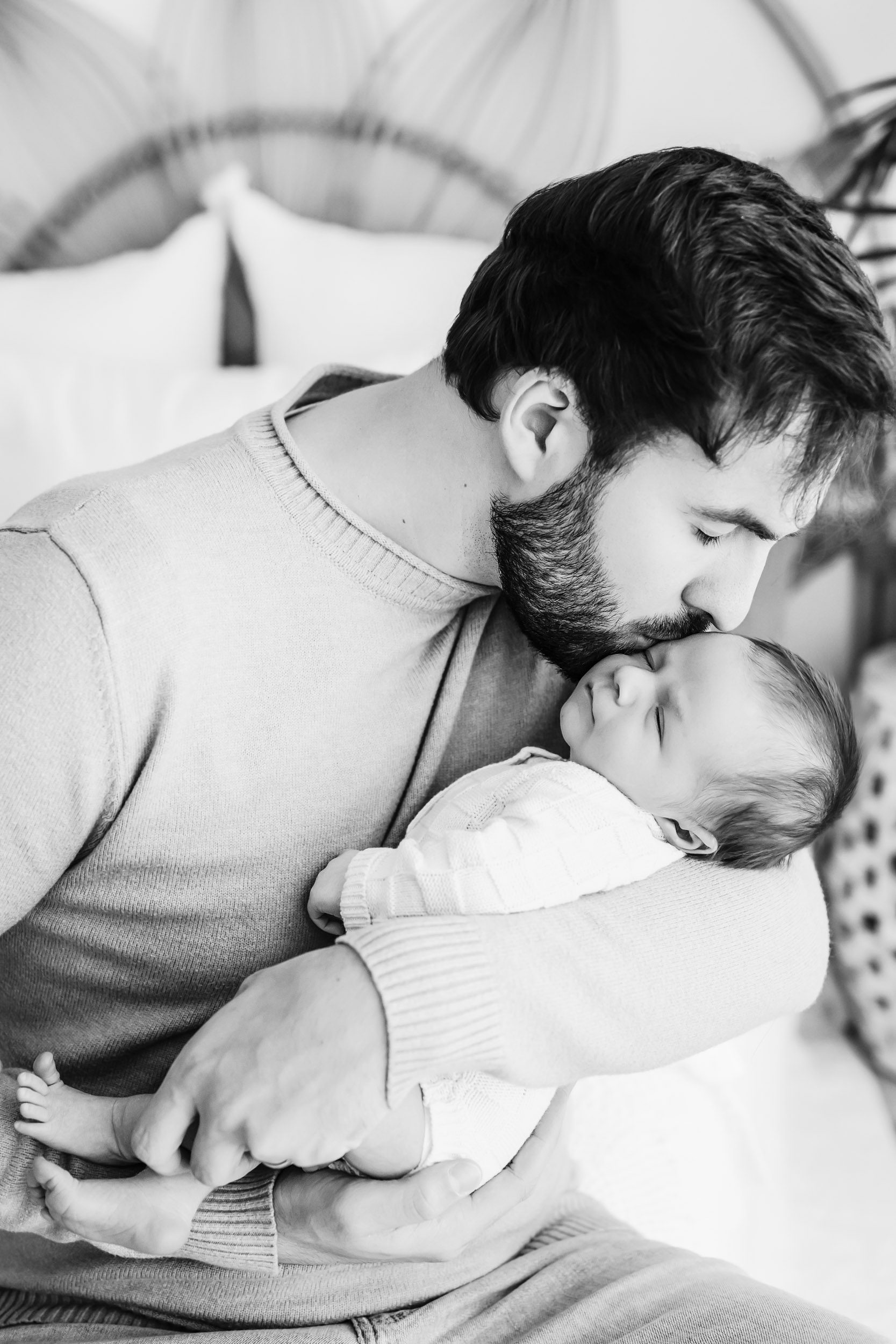 a black and white photo of a new dad sitting on a bed cradling his baby boy in his arms and giving him a gentle kiss on his temple during a newborn photography session near me