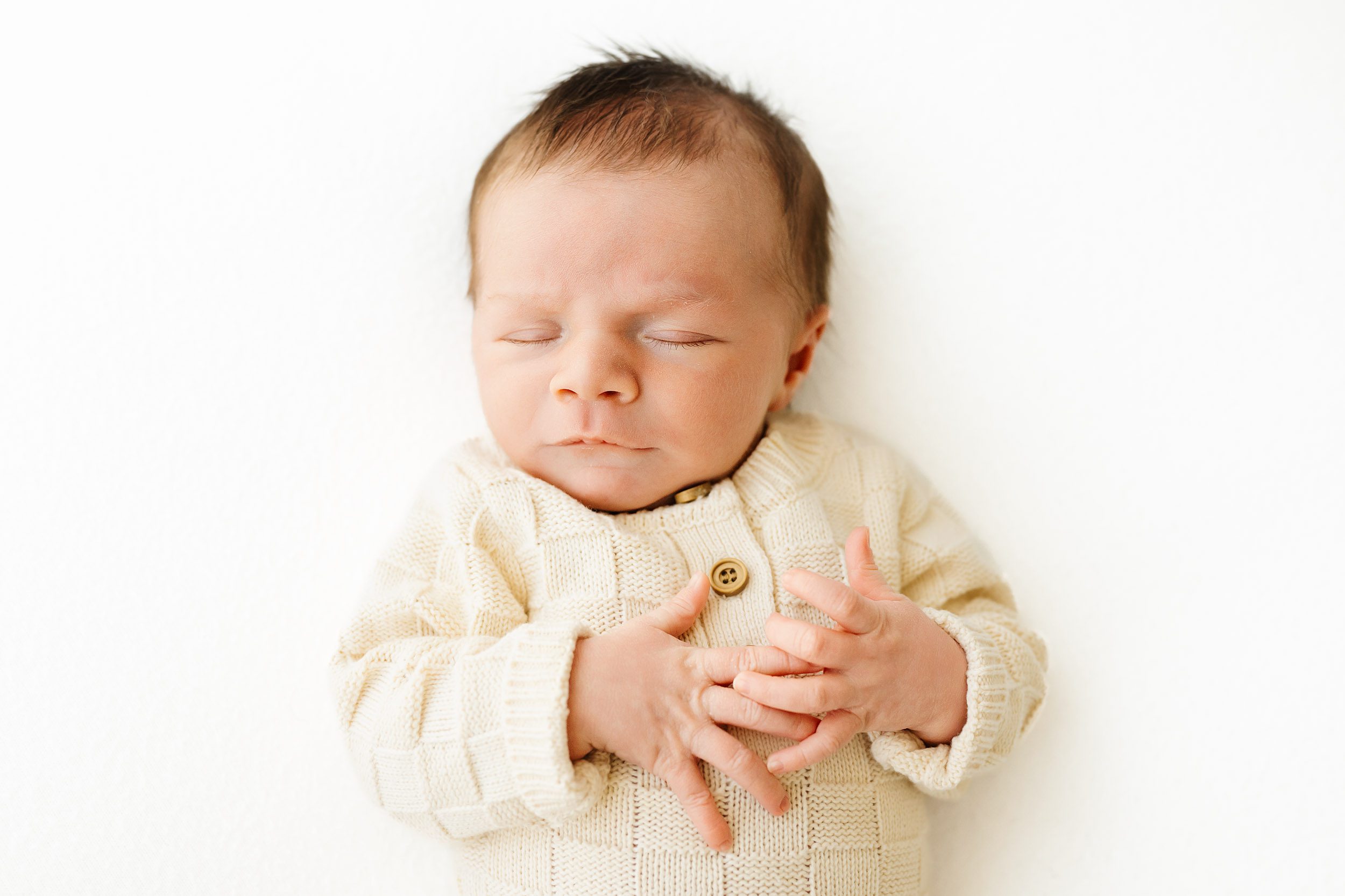 a baby boy wearing a long sleeve cream colored knit romper with a checker pattern laying on his back and sleeping peacefully during a newborn photography session near me