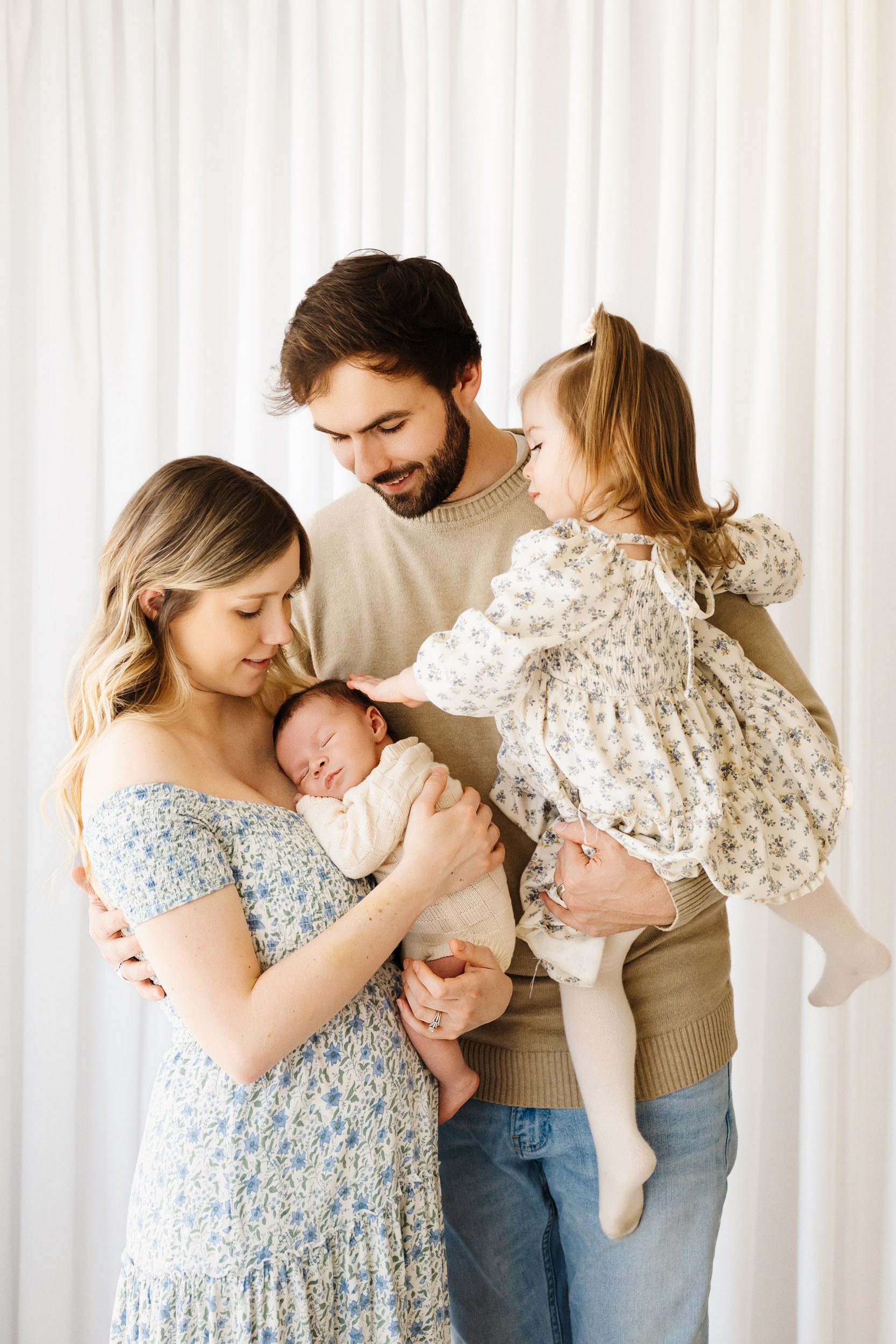a family of four standing in front of a white curtain wall while mom holds her baby boy snuggled up against her chest and dad holds big sister in his arms as she gently touches her baby brother's head during a newborn photography session near me