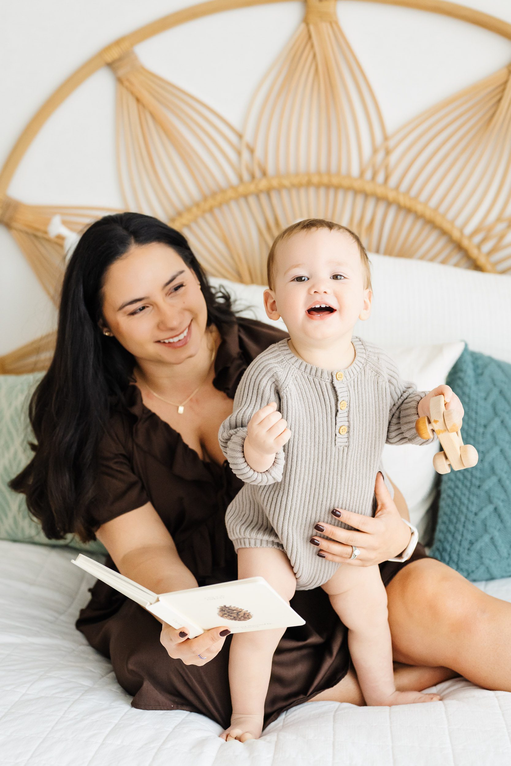 a mom sitting on a bed with her baby boy and reading him a book as he stands up and laughs at the camera during a Mommy & Me photoshoot