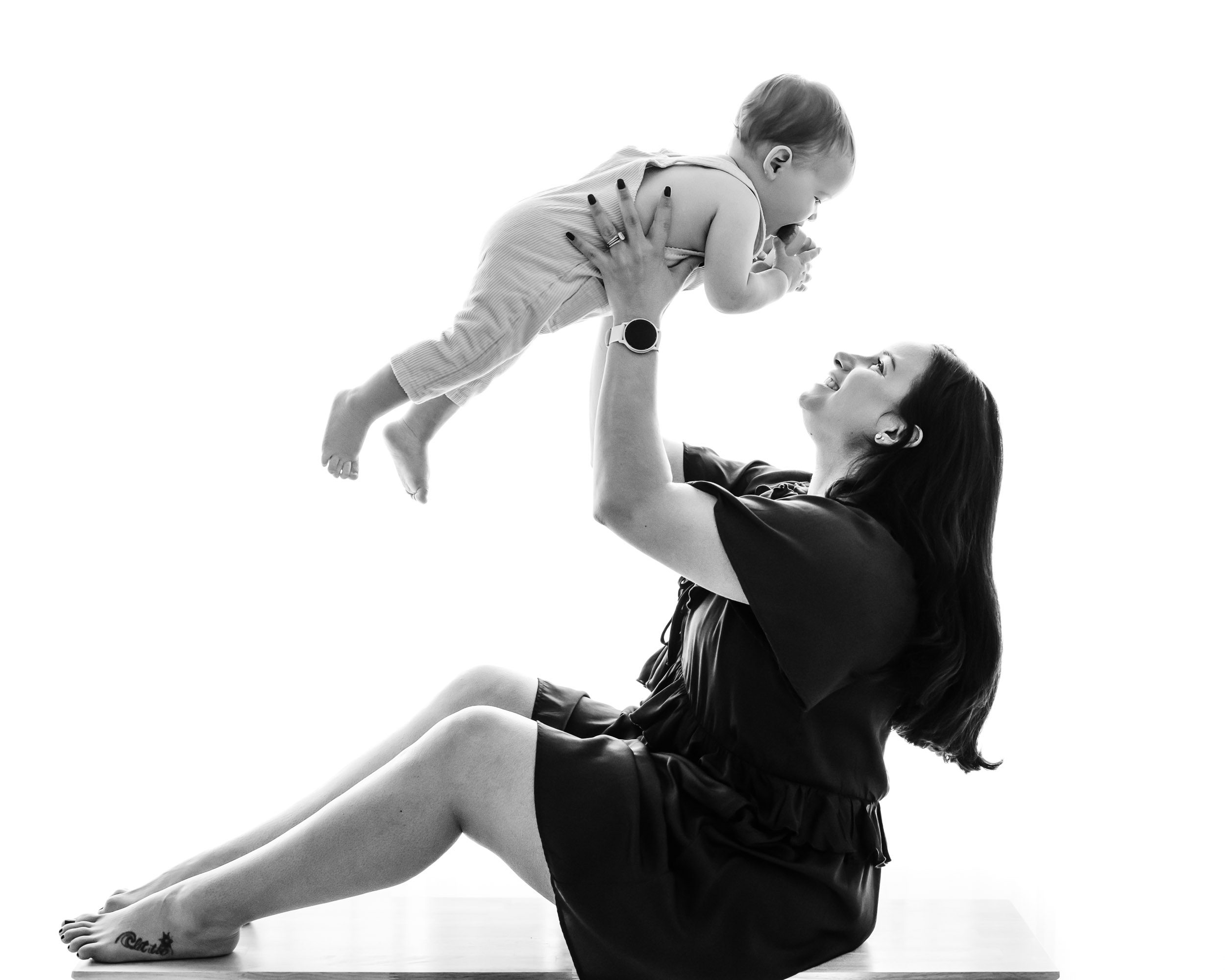 a black and white backlit photo of a mom sitting on a bench holding her young son up in the air as she smiles at him during a Mommy & Me photoshoot