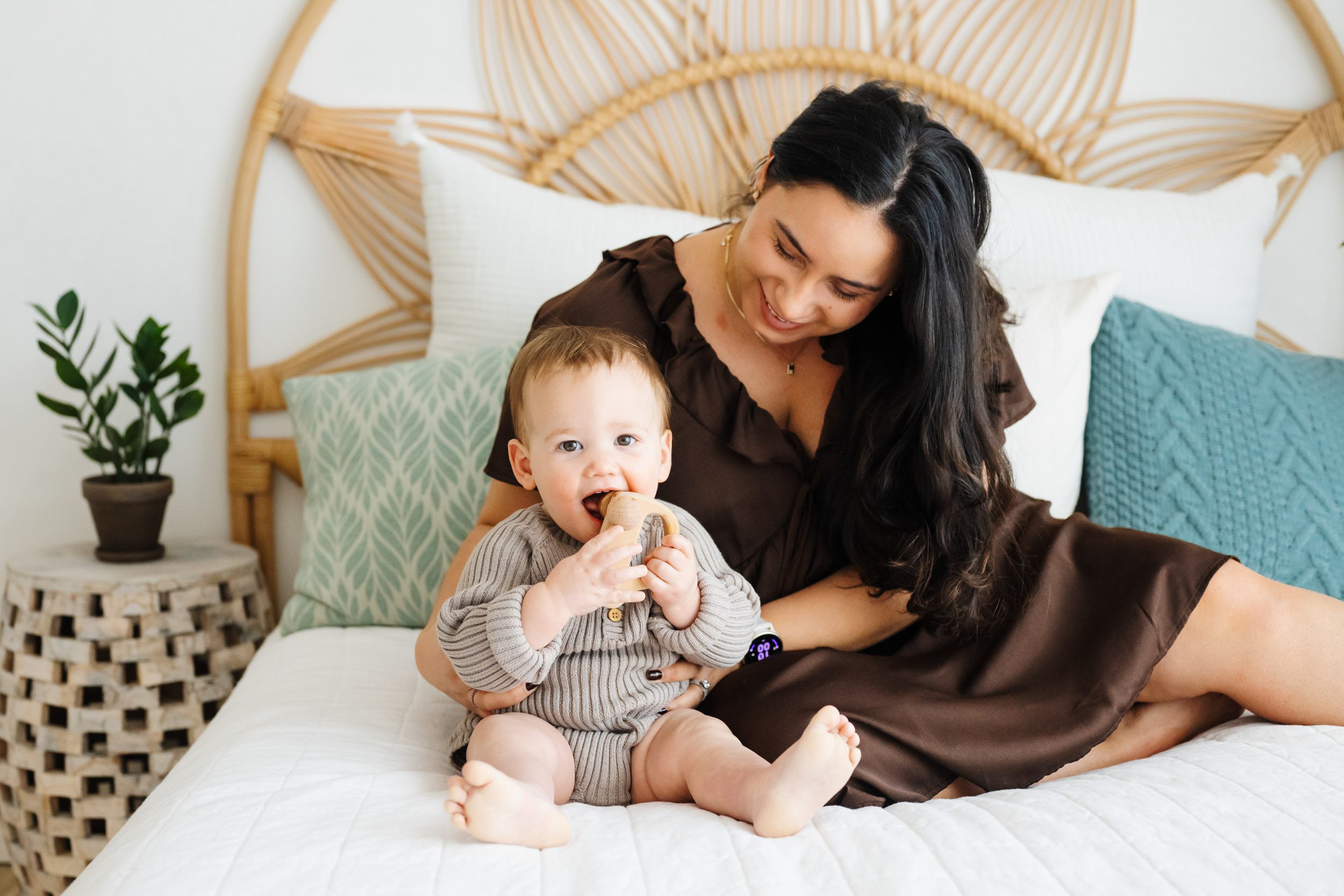 a little boy sitting on the bed putting a little wooden toy car in his mouth as his mom hugs him from behind during a Mommy & Me photoshoot