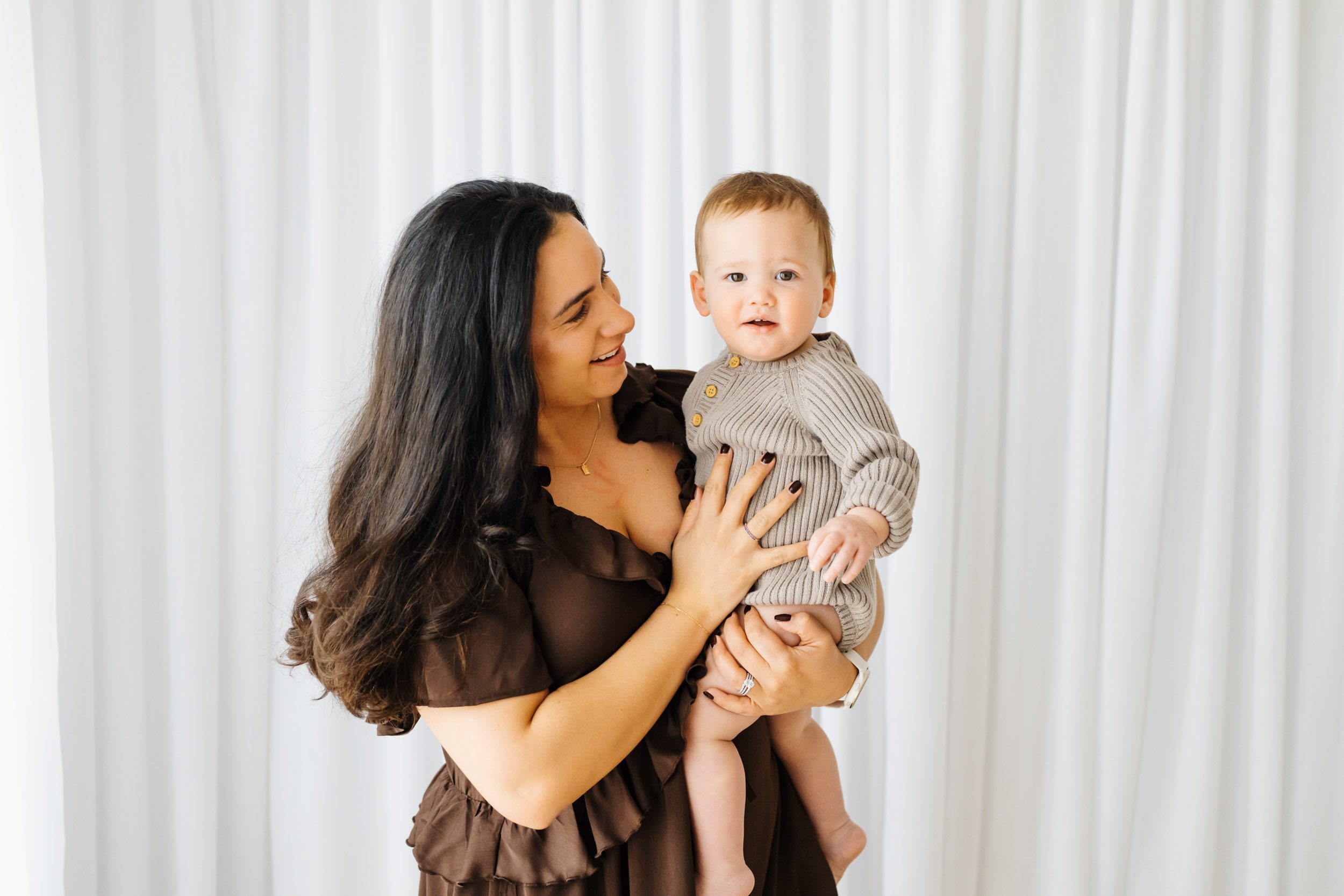a mom standing in front of a wall of white curtains holding her young son in her arms and smiling at him as he looks directly at the camera during a Mommy & Me photoshoot