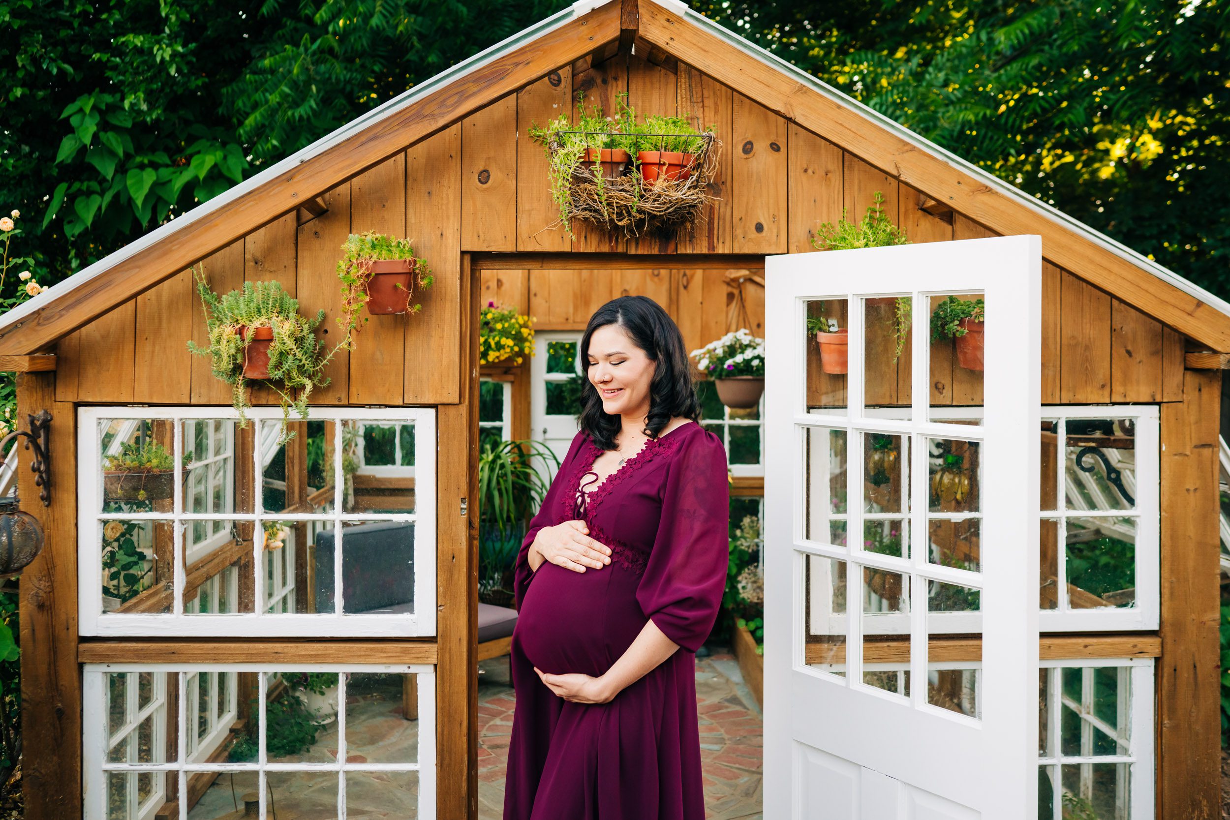 an expecting mother standing in front of a small greenhouse in a garden as she cradles her belly during a West Chester maternity photography session