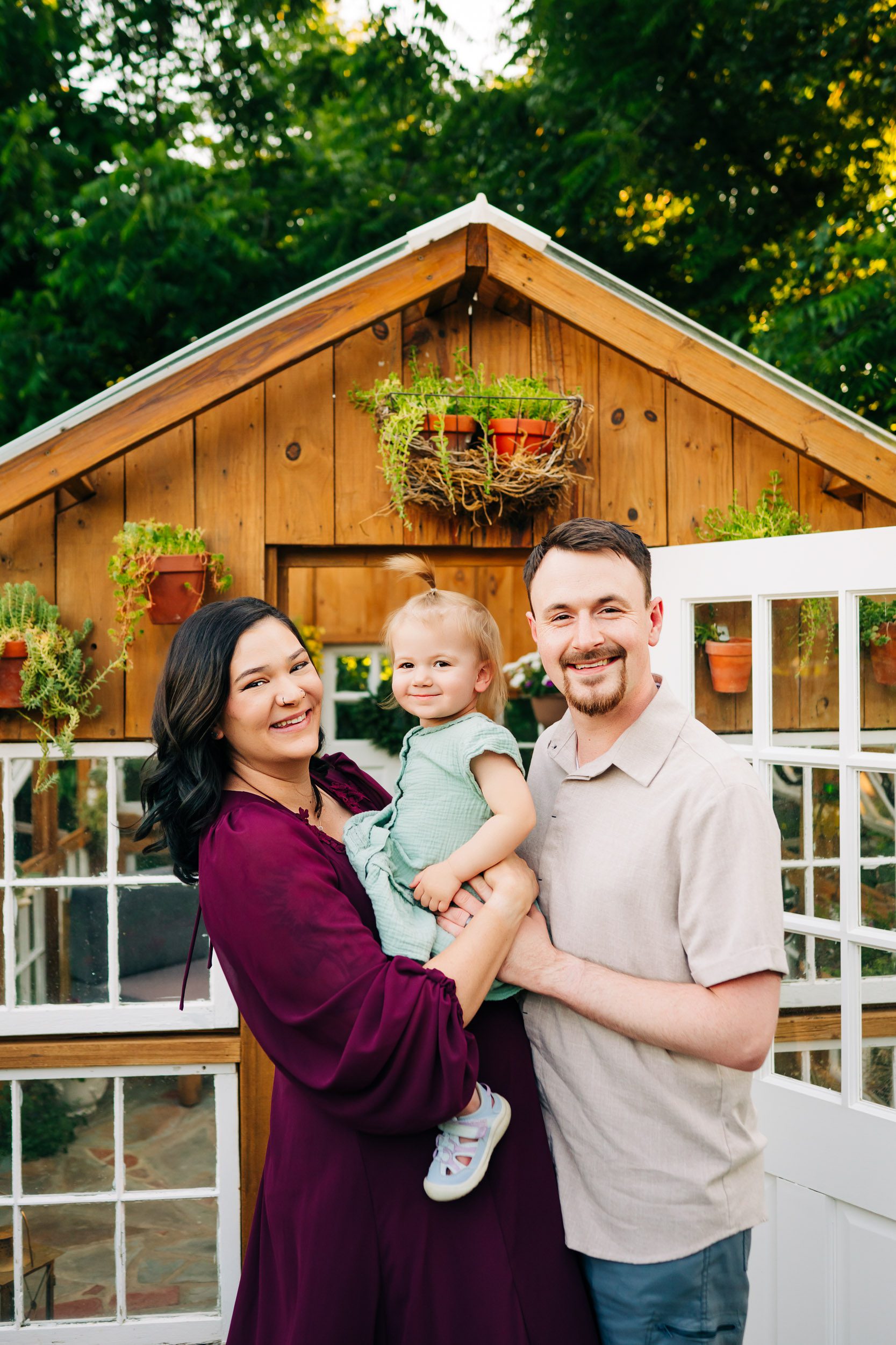 an expecting mom holding her young daughter over her belly as her partner hugs them and they all look at the camera and smile during a West Chester maternity photography session