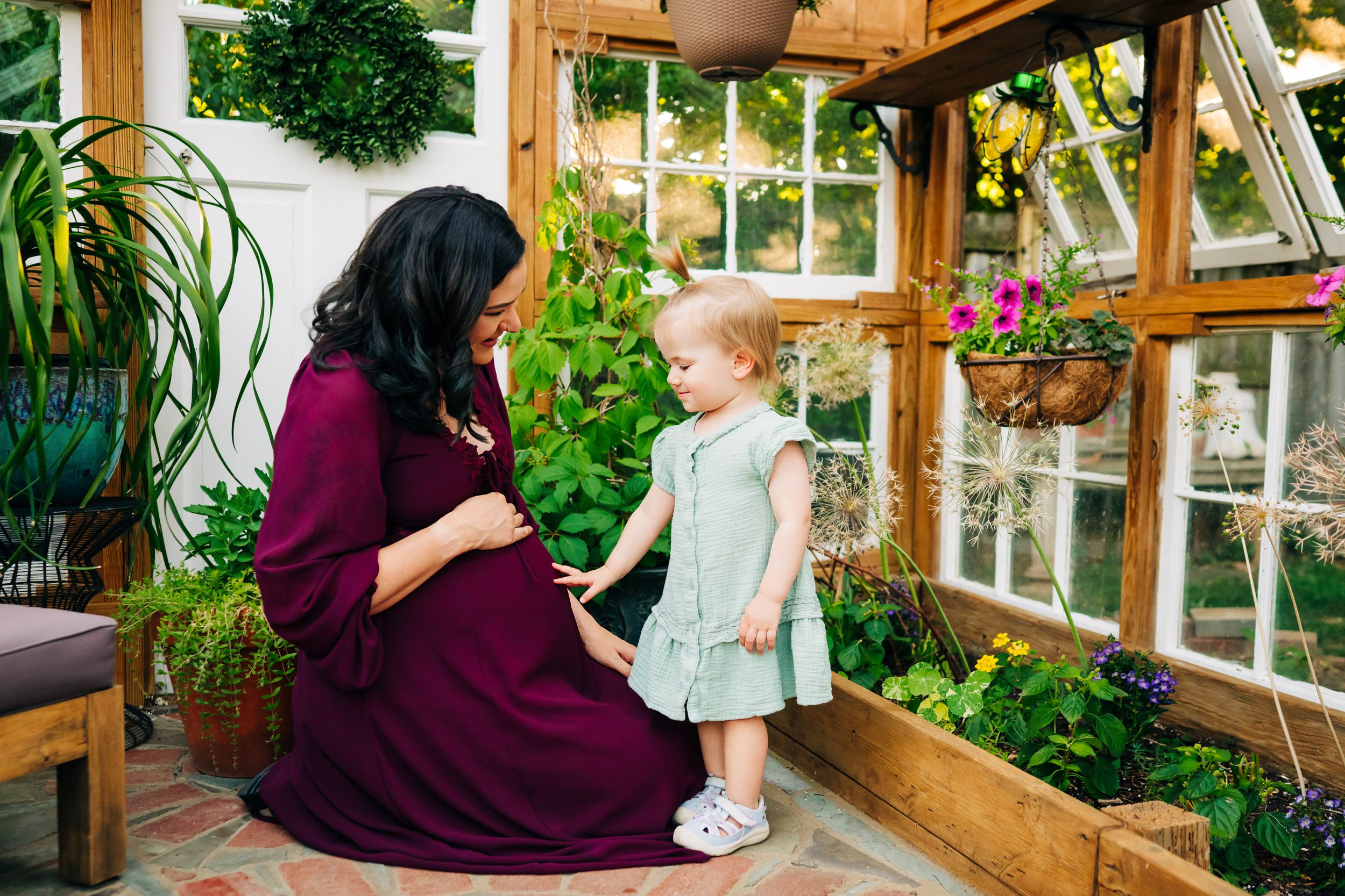a pregnant mom kneeling down in a greenhouse as her young daughter gently touches her belly during a West Chester maternity photography session