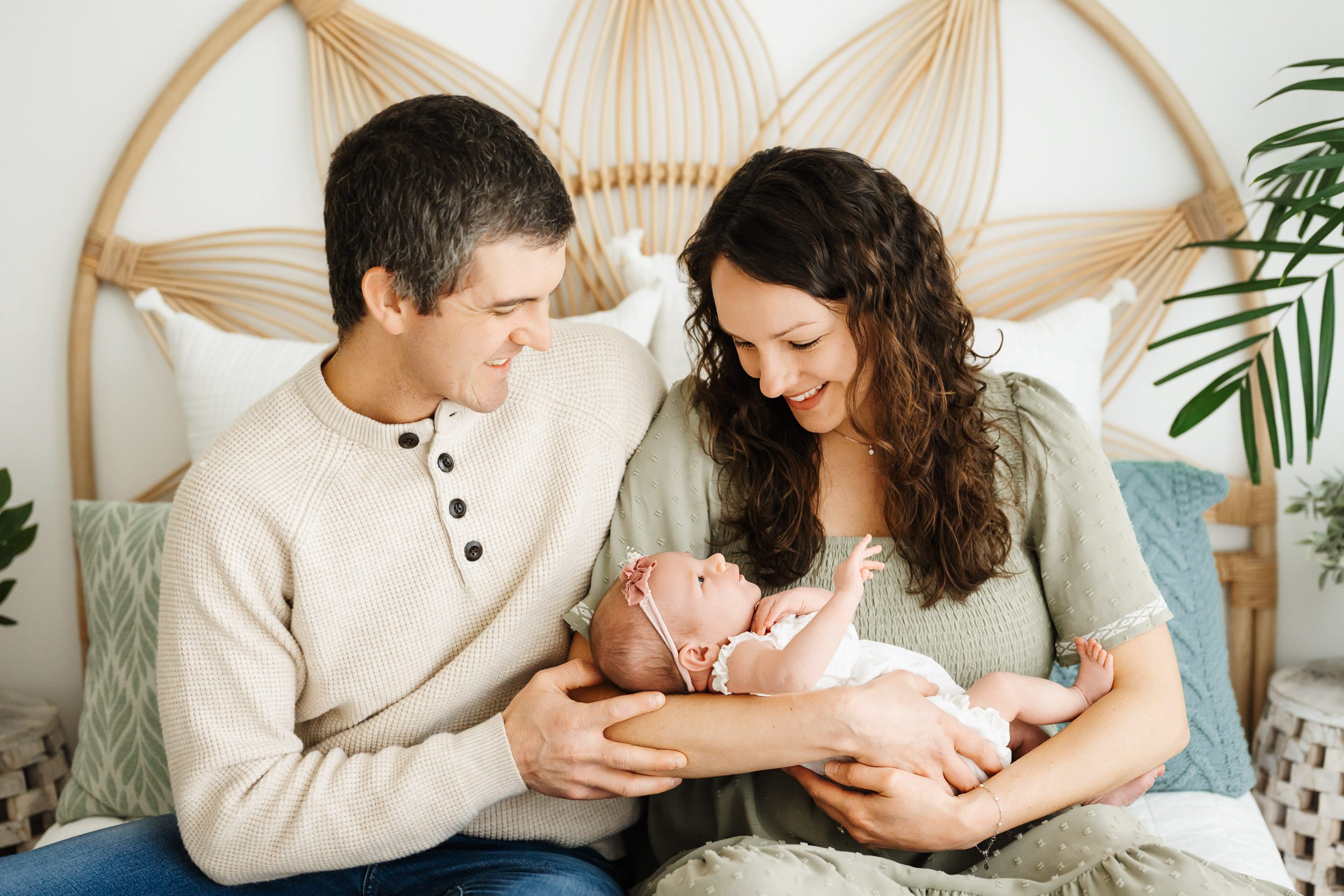 new parents sitting together on a bed cradling their baby girl in their arms and smiling down at her during a Schwenksville newborn photography session