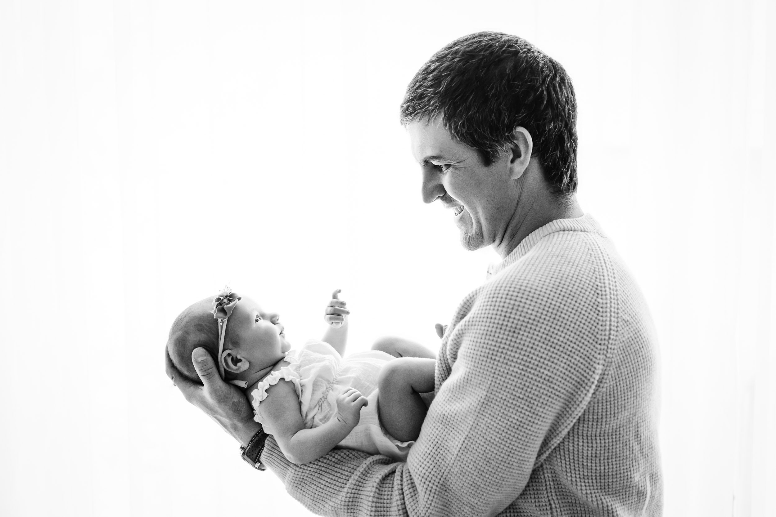 a black and white backlit photo of a new father holding his baby girl in his arms and smiling down at her during a Schwenksville newborn photography session