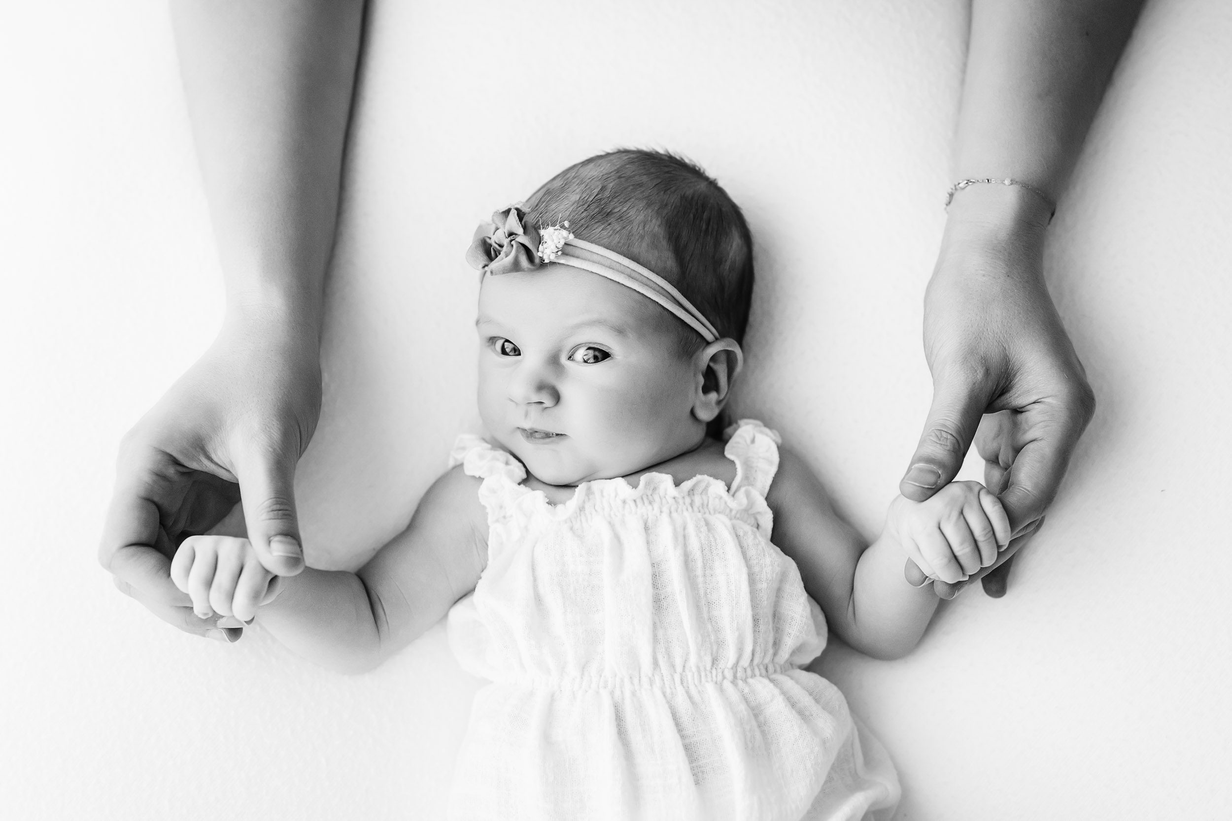 a black and white picture of a baby girl laying on a white backdrop grabbing onto her mom's fingers with her tiny hands as she gazes up at the camera during a Schwenksville newborn photography session