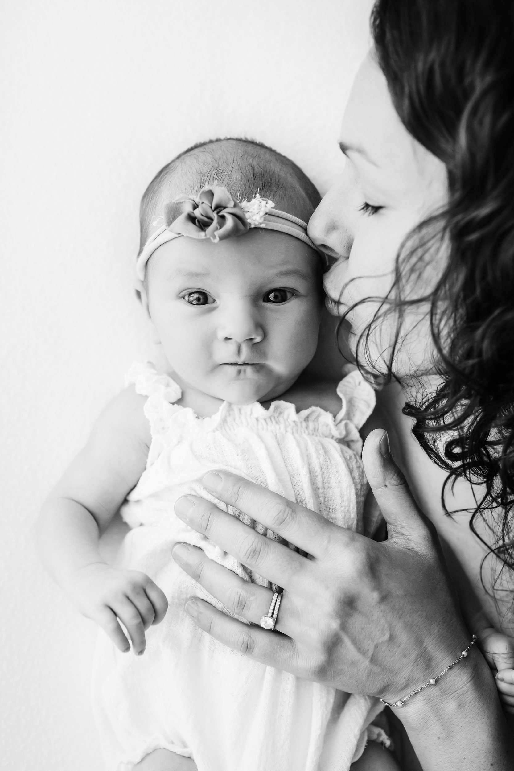 a black and white picture of a baby girl wearing a white linen romper and a headband with a bow laying on her back and gazing at the camera as her mom gently kisses her on the temple during a Schwenksville newborn photography session