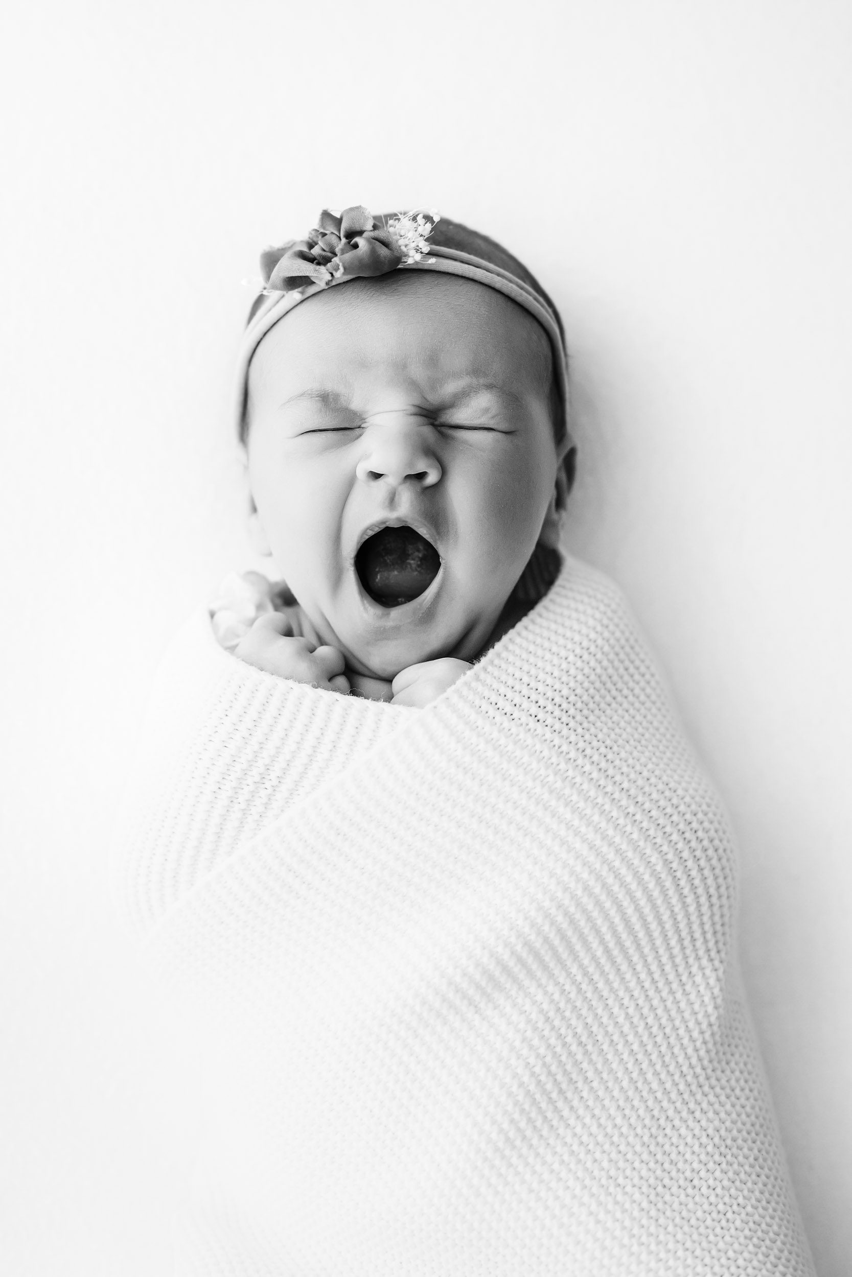 a black and white photo of a baby girl laying on her back swaddled in a textured white blanket and yawning during a Schwenksville newborn photography session