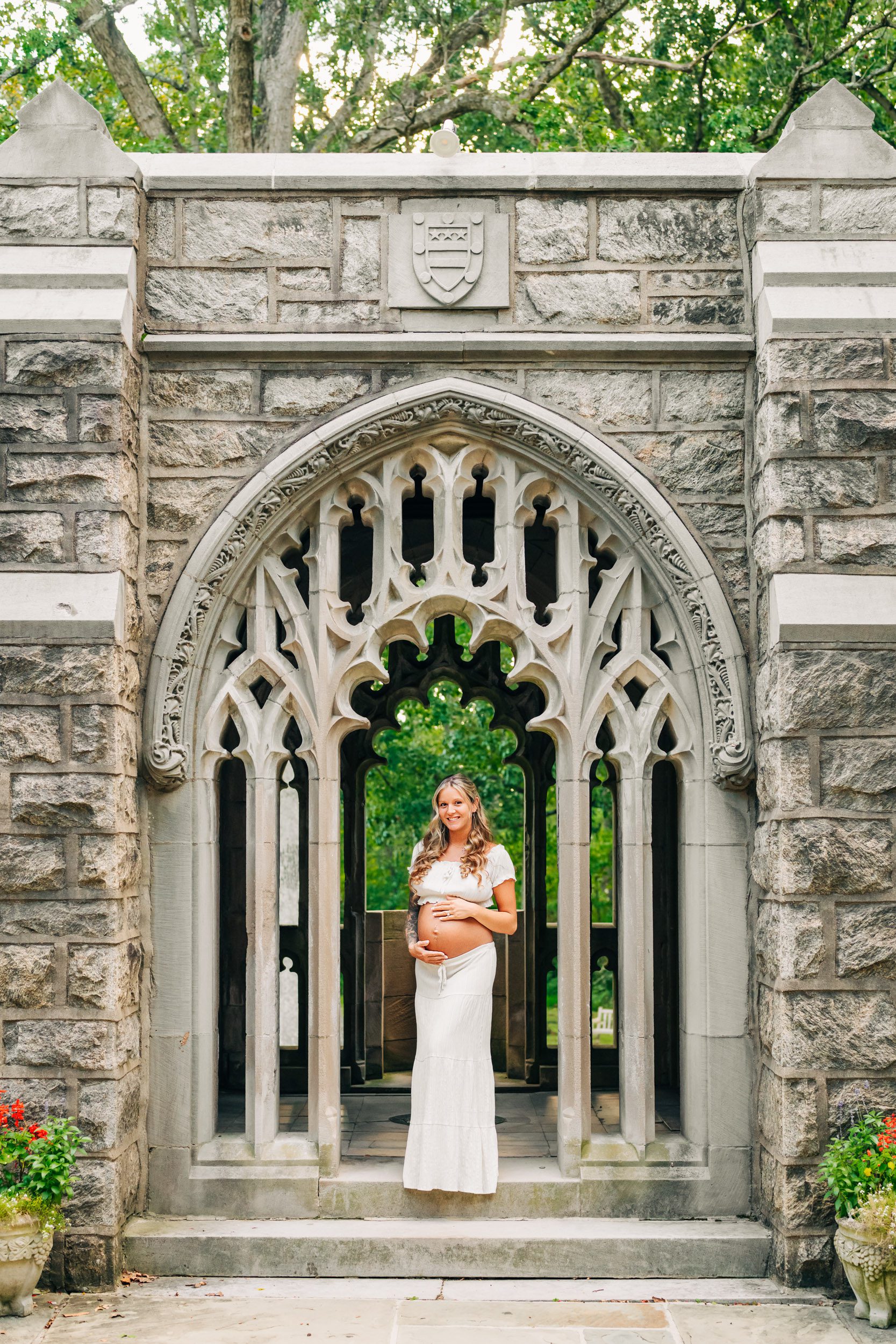an expecting mother wearing a white two piece top and skirt set and standing in a stone arch as she cradles her belly and smiles at the camera during a Valley Forge maternity photoshoot