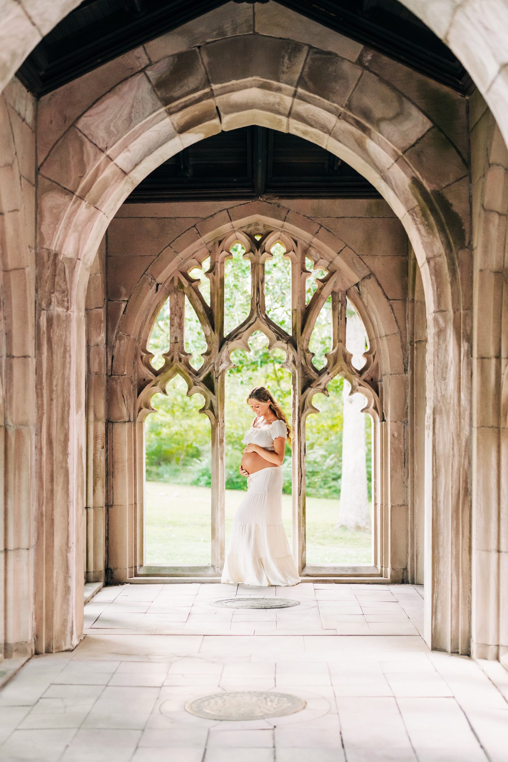 an expecting mother wearing a white two piece top and skirt set and standing in the middle of a stone arch window as she smiles down at her belly during a Valley Forge maternity photoshoot