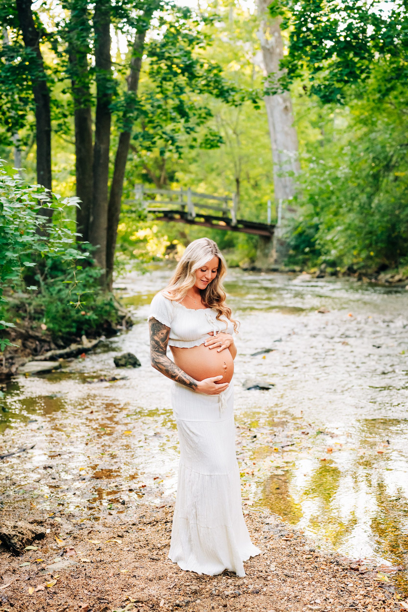 an expecting mother wearing a white two piece top and skirt set and standing next to a creek in the woods with a bridge in the background as she smiles down at her belly during a Valley Forge maternity photoshoot