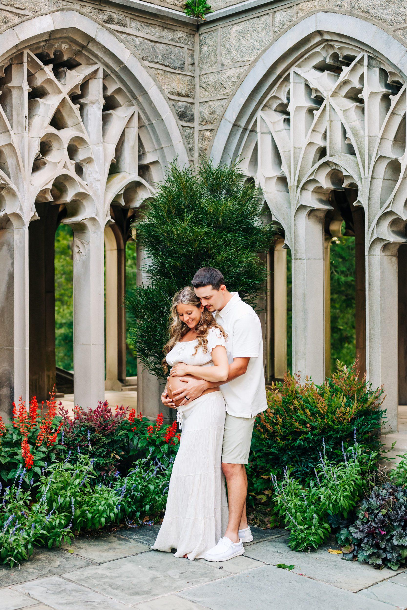 expecting parents standing in front of a small garden of flowers with stone arches in the background as dad hugs mom's belly from behind and they both smile down at her baby bump during a Valley Forge maternity photoshoot